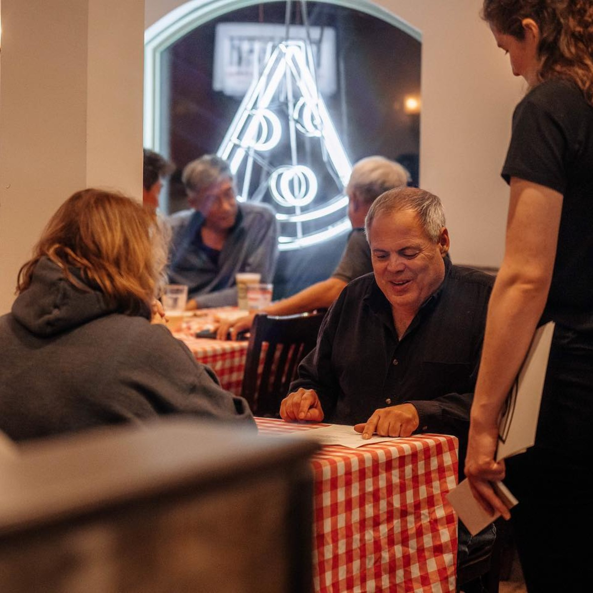 Guests at gingham tables under the neon pizza sign