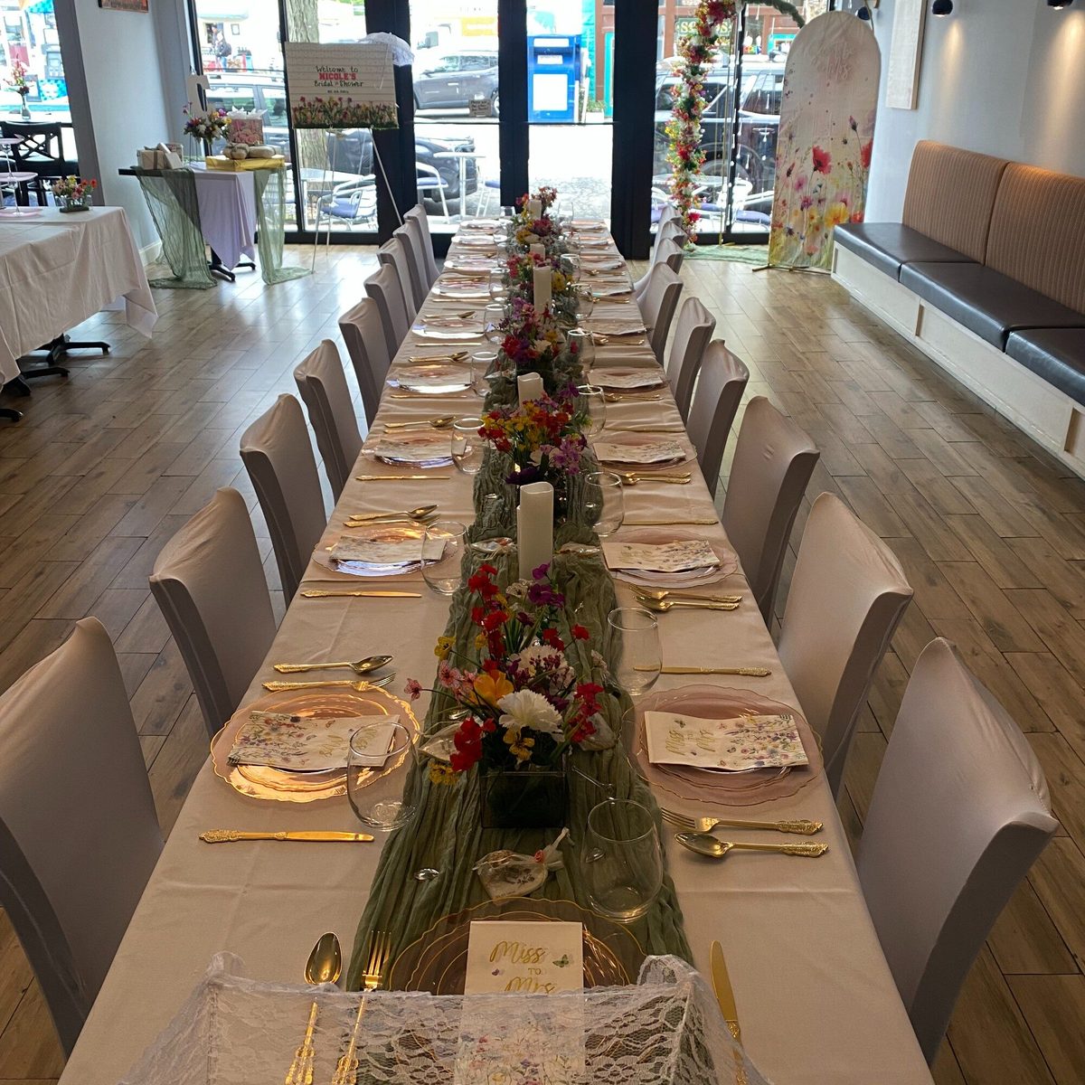 One long table down the length of Maura's Kitchen, set with white linens, gold cutlery and flowers for a bridal shower