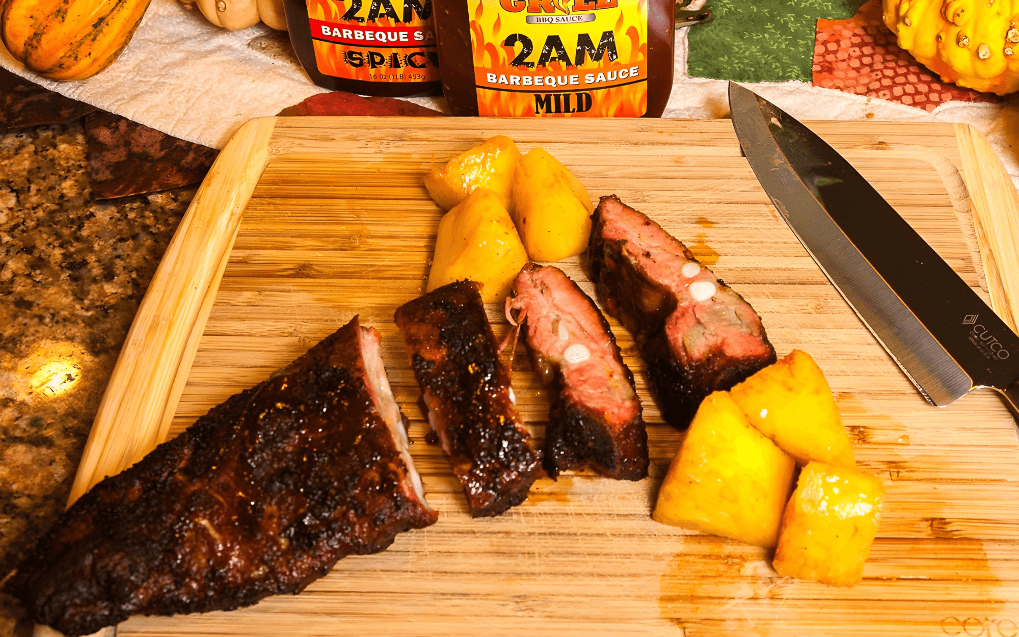 Cooked ribs and pineapple pieces on a wooden cutting board with a knife and BBQ sauce bottles in the background.