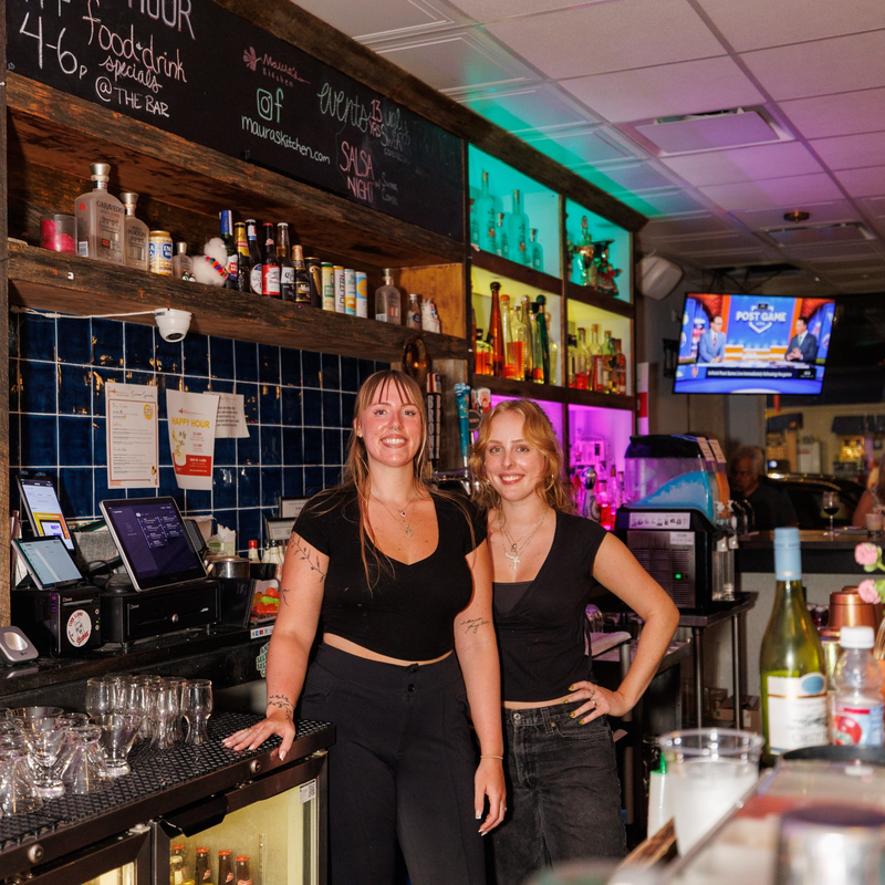 Two smiling women behind the colorful bar with a chalkboard menu